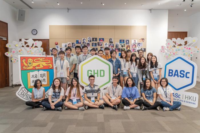 Students sitting in front or standing behind three signs showing HKU crest, GHD, and BASC
