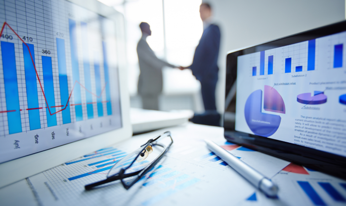 Image of eyeglasses, pen, two touchpads and financial documents at workplace with businessmen handshaking on background