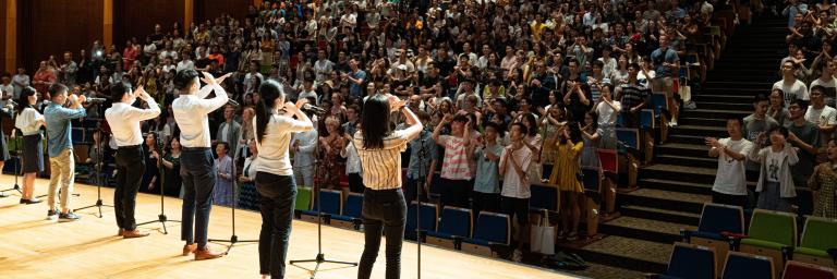 Students making a cross with their arms on a stage in a large lecture hall full of people